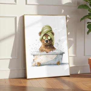 以下が含まれることがあります： ヴィンテージのバスタブでリラックスする茶色のクマが描かれた、額入りのアートプリント。クマは緑色のタオルとキュウリのスライスを目の上に着用しています。小さなボトルが浴槽の横にあります。アートワークは金色のフレームに入っています。