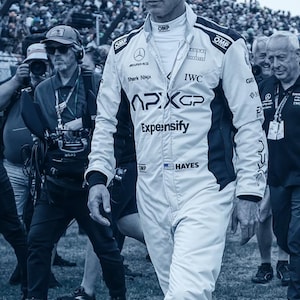 May include: A person wearing a white racing suit with black accents, the suit features logos for APXGP, IWC, and Expensify. The person is walking through a crowd of people, with a blurred background of spectators and a stadium.