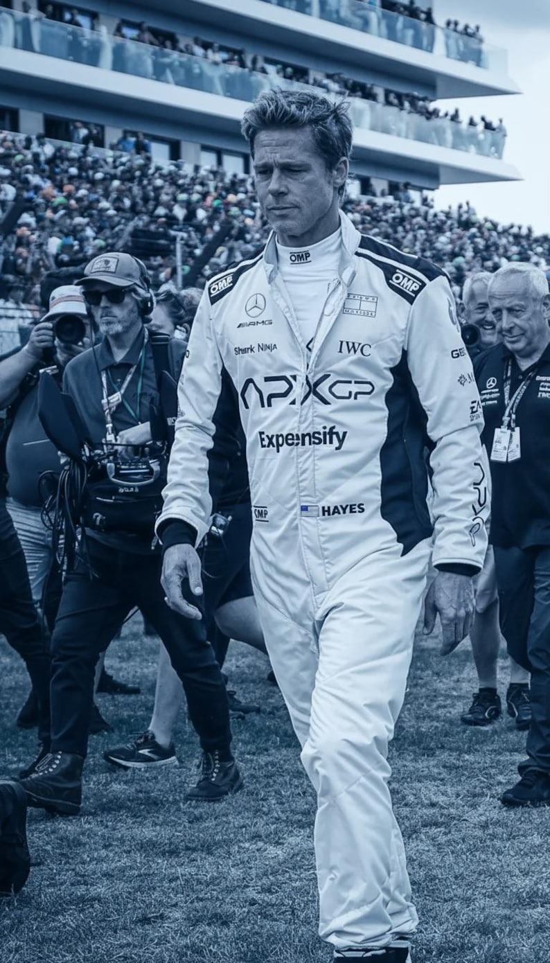 May include: A person wearing a white racing suit with black accents, the suit features logos for APXGP, IWC, and Expensify. The person is walking through a crowd of people, with a blurred background of spectators and a stadium.