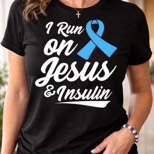 Könnte beinhalten: Schwarzes T-Shirt mit dem Text "I Run on Jesus & Insulin" in weißer Schrift und einer hellblauen Schleifengrafik. Das Shirt hat einen Rundhalsausschnitt und kurze Ärmel.
