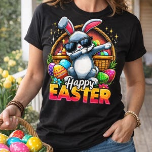 Puede incluir: Camiseta negra con un gráfico colorido de "Felices Pascuas". El gráfico presenta un conejito gris con gafas de sol, haciendo un dab junto a una cesta de huevos de Pascua. Una cesta de mimbre llena de coloridos huevos de Pascua está en primer plano.