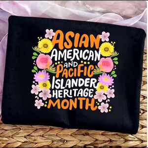 Könnte beinhalten: Schwarzes T-Shirt mit dem Text "ASIAN AMERICAN AND PACIFIC ISLANDER HERITAGE MONTH" in Orange und Weiß. Der Text ist von bunten Blumenillustrationen in Rosa, Gelb und Grün umgeben.