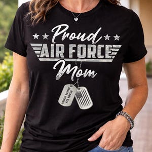 Op de afbeelding: Zwart t-shirt met de witte tekst "Proud Air Force Mom". Het ontwerp bevat sterren en hondenpenningen met de Amerikaanse vlag en de tekst "United States Air Force".