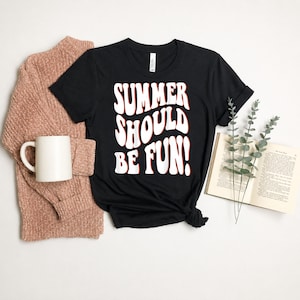 Może przedstawiać: Czarny t-shirt z napisem "SUMMER SHOULD BE FUN!" w kolorze białym z czerwonym obrysem. Różowy sweter, biały kubek i otwarta książka również znajdują się na zdjęciu. Koszulka to casualowy element letniej odzieży.