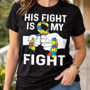 May include: Black t-shirt with the text "HIS FIGHT IS MY FIGHT" in white. The shirt features a colorful puzzle ribbon design held by two white hands. The ribbon is a symbol of autism awareness.