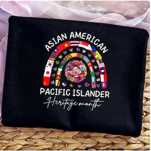 Puede incluir: Bolso tote negro con un diseño de arcoíris con banderas y flores, con el texto "ASIAN AMERICAN PACIFIC ISLANDER Heritage month" en blanco. El bolso se muestra sobre una superficie tejida.