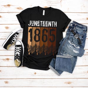 May include: Black t-shirt with the word "JUNETEENTH" in white above the number "1865" in black. The number is designed with a gradient of skin tones. The shirt is paired with black sneakers and blue jeans.