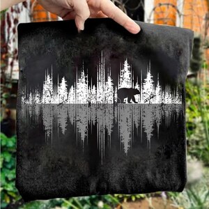 May include: Black square pillow with a white forest silhouette design and a bear walking across the trees. The design is mirrored below the trees. The pillow is held by a hand.