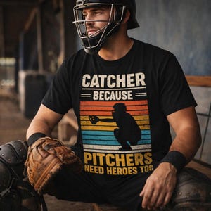 May include: Black t-shirt with the text "CATCHER BECAUSE PITCHERS NEED HEROES TOO" in white. The shirt features a silhouette of a catcher in front of a retro sunset design. The person is wearing a catcher's helmet and gear.