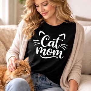 以下が含まれることがあります： 白い「Cat mom」の文字と猫の耳のグラフィックが入った黒いTシャツ。青いジーンズとベージュのカーディガンを着用。オレンジ色の猫が人の膝の上に座っています。