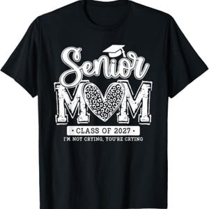 May include: A black short-sleeve t-shirt featuring white graphic text that reads Senior MOM with a leopard print heart replacing the letter O. Above the text is a graduation cap. Below is the text CLASS OF 2027 and I'M NOT CRYING, YOU'RE CRYING.