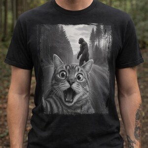 May include: Black t-shirt featuring a grayscale image of a surprised cat on a road, with a Bigfoot figure in the background. The cat has wide eyes and an open mouth. The background shows a forest scene.