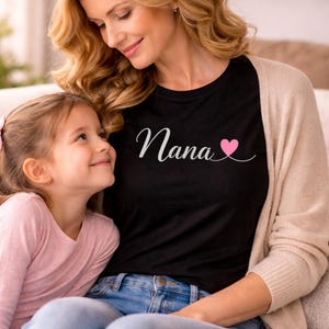 May include: Black raglan sleeve shirt with the word "Nana" in white script and a pink heart. A woman and a young girl are looking at each other. The woman is wearing a beige cardigan and blue jeans.