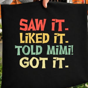 May include: Black tote bag featuring the text "SAW IT. LIKED IT. TOLD MIMI! GOT IT." in red, yellow, green, and yellow block letters. The bag is held up by a person.