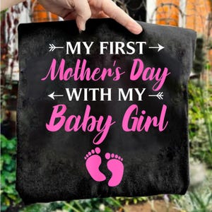 May include: Black sign with the text "MY FIRST Mother's Day WITH MY Baby Girl" in white and pink. Pink baby footprints are at the bottom. The sign is held up by a hand.