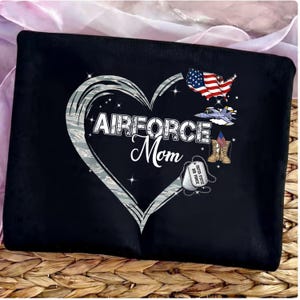 May include: Black tote bag featuring a heart-shaped design with the words "AIRFORCE Mom" in white. The heart is outlined in gray and filled with a camouflage pattern. Includes an American flag, a jet, dog tags, and boots.