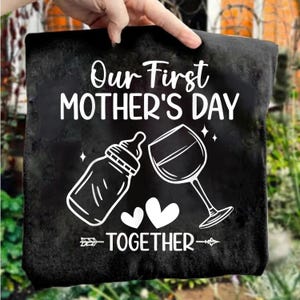 May include: Black square decorative pillow with white text that reads "Our First MOTHER'S DAY TOGETHER." The design includes a baby bottle, a wine glass, two hearts, and an arrow.