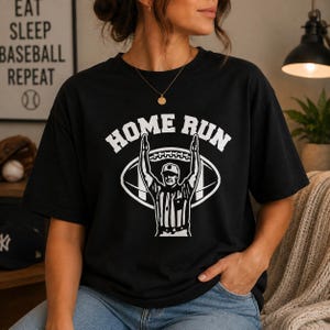 May include: A black short-sleeve t-shirt featuring a white graphic print of a sports referee with arms raised in a touchdown signal. The text HOME RUN is arched above the referee, and a football shape is positioned behind the figure.
