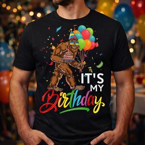Puede incluir: Camiseta negra con un diseño de Bigfoot de dibujos animados sosteniendo un pastel y globos. El texto "IT'S MY Birthday" está en blanco y colores arcoíris. Confeti añade al tema de cumpleaños, creando un diseño festivo.