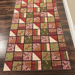 May include: A rectangular quilted table runner featuring a patchwork design of squares and triangles. The fabric patterns include olive green, deep red, and cream colors with dragonfly motifs, floral prints, and swirling line designs.