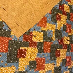 Pu&ograve; includere: Un top per trapunta patchwork con vari quadrati di tessuto in tonalit&agrave; giallo senape, verde oliva, arancione ruggine e azzurro con motivi floreali e a pois, parzialmente coperto da un tessuto giallo senape con spille da balia.