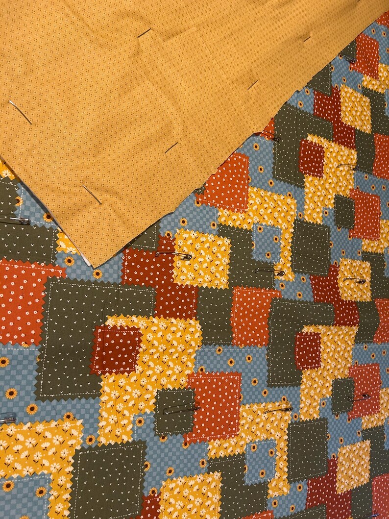 Pu&ograve; includere: Un top per trapunta patchwork con vari quadrati di tessuto in tonalit&agrave; giallo senape, verde oliva, arancione ruggine e azzurro con motivi floreali e a pois, parzialmente coperto da un tessuto giallo senape con spille da balia.