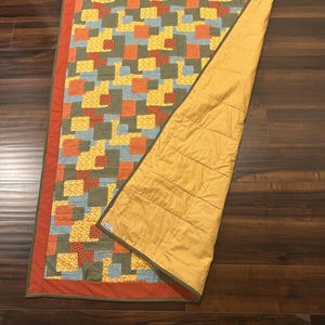 Pu&ograve; includere: Una trapunta rettangolare con un motivo patchwork di quadrati gialli, arancioni, blu e verde oliva con un bordo arancione a tinta unita. Il retro &egrave; in tessuto giallo senape a tinta unita con linee di cucitura orizzontali visibili.