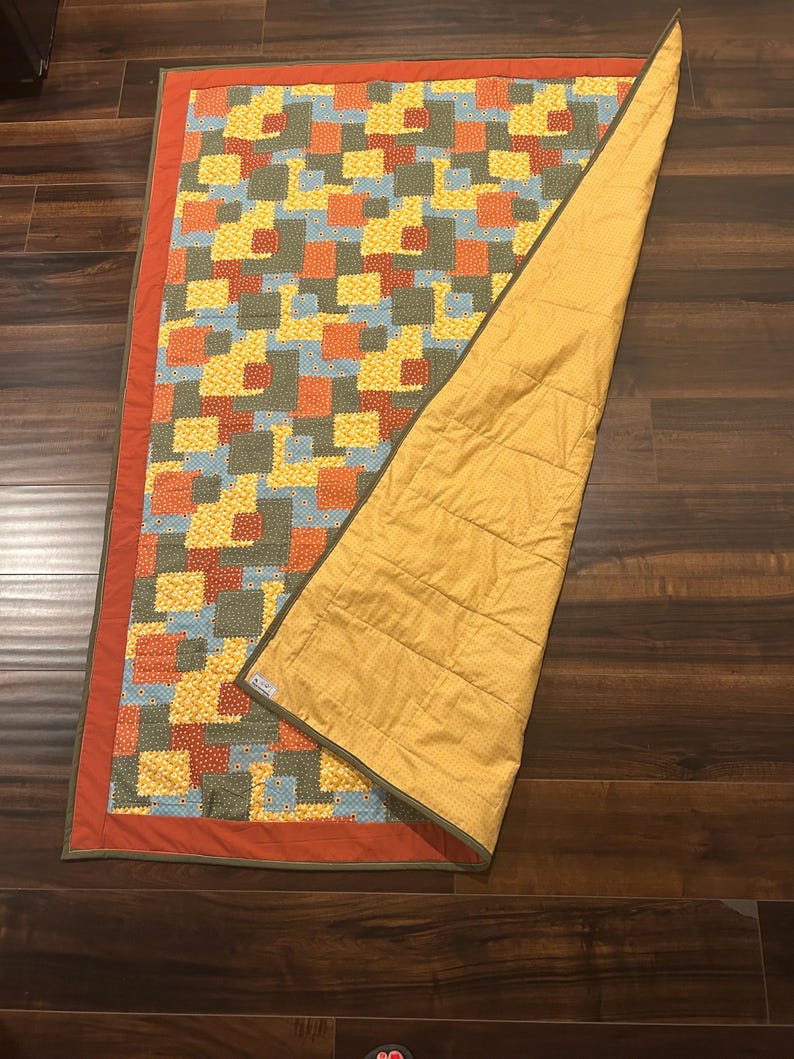 Pu&ograve; includere: Una trapunta rettangolare con un motivo patchwork di quadrati gialli, arancioni, blu e verde oliva con un bordo arancione a tinta unita. Il retro &egrave; in tessuto giallo senape a tinta unita con linee di cucitura orizzontali visibili.