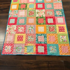 May include: A rectangular patchwork quilt featuring a grid of square blocks in various colors including red, orange, yellow, green, blue, and pink. The quilt displays a mix of floral, polka dot, plaid, and geometric patterns with a bright blue border.