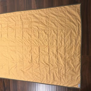 Pu&ograve; includere: Una trapunta rettangolare in tessuto giallo senape con un sottile motivo a griglia. La trapunta &egrave; rifinita con un bordo verde oliva scuro lungo tutti e quattro i lati e presenta una griglia di linee di cucitura verticali e orizzontali.