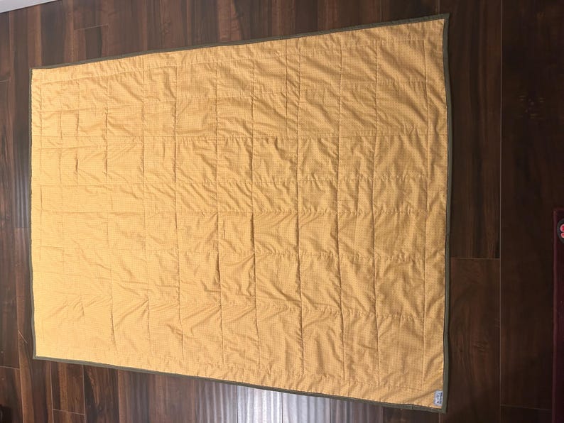 Pu&ograve; includere: Una trapunta rettangolare in tessuto giallo senape con un sottile motivo a griglia. La trapunta &egrave; rifinita con un bordo verde oliva scuro lungo tutti e quattro i lati e presenta una griglia di linee di cucitura verticali e orizzontali.