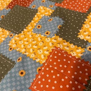 Pu&ograve; includere: Una trapunta patchwork con pezzi di tessuto rettangolari dai bordi dentellati. Il motivo include verde oliva a pois bianchi, arancione bruciato con fiori bianchi, quadretti vichy azzurri e giallo brillante con stampe floreali a margherita.