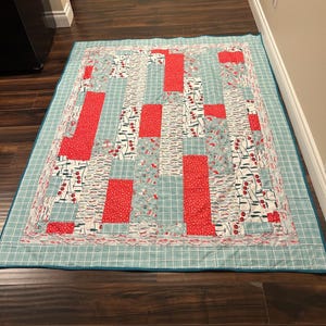 Può includere: Una trapunta fatta a mano con un design patchwork. La trapunta presenta blocchi rettangolari rossi e sezioni di tessuto fantasia con un motivo a griglia blu chiaro e bianco. Il bordo è blu chiaro unito.
