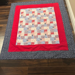 Może przedstawiać: Ręcznie robiona kołdra o tematyce patriotycznej. Kołdra posiada centralny panel z wzorami flag w kolorach czerwonym, białym i niebieskim, obramowany czerwoną obwódką i granatową zewnętrzną obwódką z białymi kropkami. Kołdra jest prawdopodobnie wykonana z bawełny.