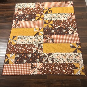 May include: A rectangular patchwork quilt featuring a repeating pattern of mustard yellow, orange gingham, and dark brown fabric panels decorated with white floral and leaf motifs, accented by small pinwheel blocks throughout the design.