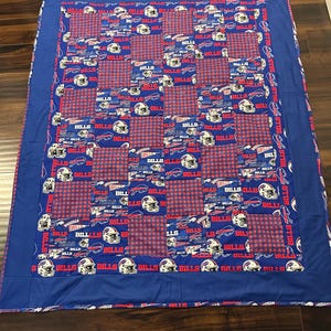 May include: A rectangular quilt featuring a patchwork design of Buffalo Bills football team fabric. The pattern alternates between squares of blue fabric printed with white helmets and red text, and squares of a red and blue checkered fabric pattern.