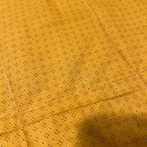 Pu&ograve; includere: Un tessuto giallo senape con un motivo ripetuto di minuscole stelle a quattro punte marrone scuro. Il tessuto presenta cuciture dritte visibili che creano un motivo a griglia sulla superficie del materiale.