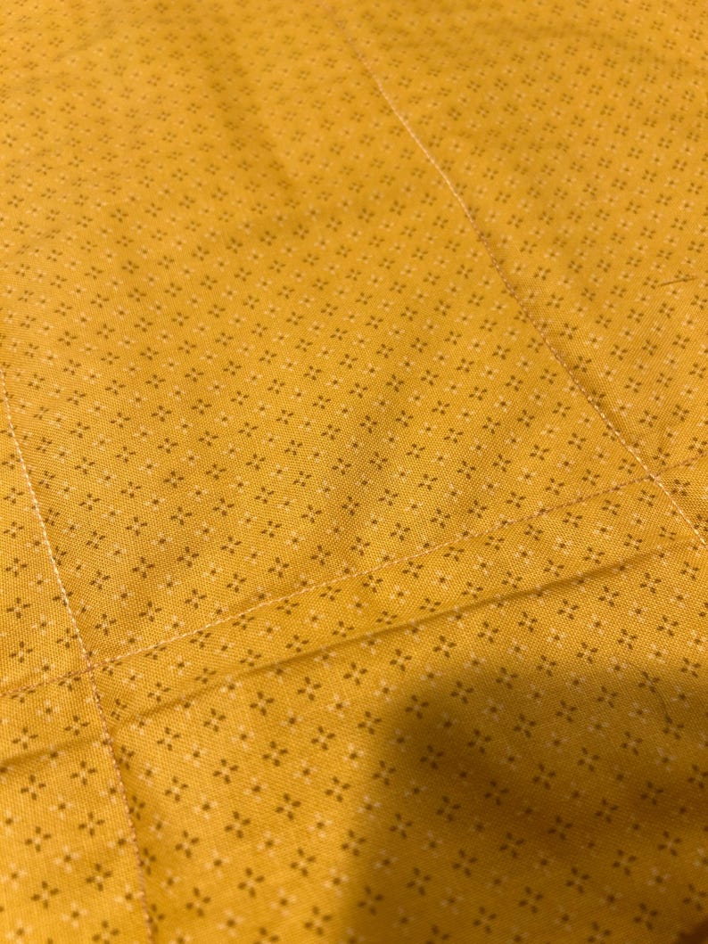 Pu&ograve; includere: Un tessuto giallo senape con un motivo ripetuto di minuscole stelle a quattro punte marrone scuro. Il tessuto presenta cuciture dritte visibili che creano un motivo a griglia sulla superficie del materiale.