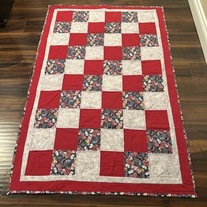 Op de afbeelding: Een handgemaakte quilt met een rood en wit dambordpatroon. De vierkanten wisselen af tussen effen rood, wit en een patriottische print. De quilt heeft een rode rand en is geschikt voor beddengoed of decoratie.
