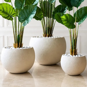 May include: Three artificial plants with large, vibrant green leaves housed in spherical, off-white textured planters. Each pot is filled with small, smooth white stones at the base of the stems, creating a uniform look across the three different sizes.