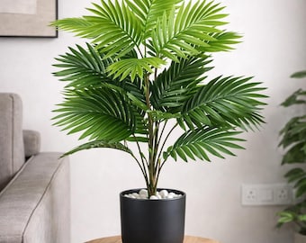 Op de afbeelding: Een heldergroene kunstpalm in een zwarte pot gevuld met witte stenen. De plant staat op een ronde houten tafel met zwarte metalen poten. De achtergrond omvat een grijze bank en een ingelijst kunstwerk.