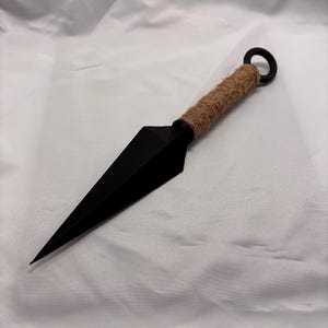 May include: A black kunai knife with a sharp, triangular blade. The handle is wrapped in brown twine, and a black ring is at the end. The knife is displayed against a white background.