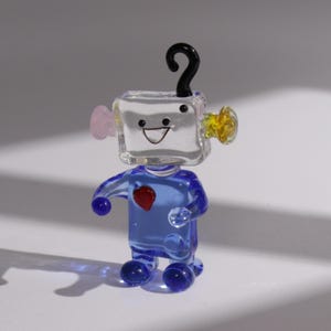 以下が含まれることがあります： 風変わりなガラス製のロボットの置物。ロボットは青い体、笑顔の顔の透明な四角い頭、そして上部に黒い疑問符が付いています。ピンクと黄色の耳のようなアクセントと、胸に赤いハートが付いています。