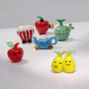 以下が含まれることがあります： 果物、ポップコーン、ティーポットの形をした、気まぐれなガラス製の置物のコレクション。 赤いリンゴ、黄色い梨、青いティーポットなど、鮮やかな色をしています。 各置物には笑顔があります。