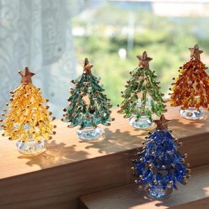 May include: Five miniature glass Christmas trees in shades of gold, green, and blue, each topped with a star. The trees have a textured, beaded appearance and sit on clear, round bases, displayed on a wooden surface.