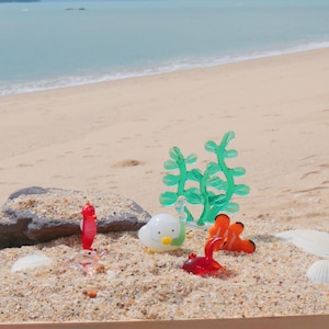 Peut inclure: Une collection de créatures et de plantes marines en verre coloré sur une surface sablonneuse. Les figurines comprennent un hippocampe rouge, un escargot blanc avec un bec jaune, deux poissons-clowns oranges et un crabe rouge. Des plantes vertes ressemblant à des algues sont également présentes.