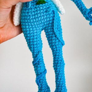 May include: A handmade crochet doll featuring a bright blue body with textured stitching. The doll has white wing-like appendages attached to the upper back and a small green accent detail at the waist. The legs are long and slender with blue yarn detailing.