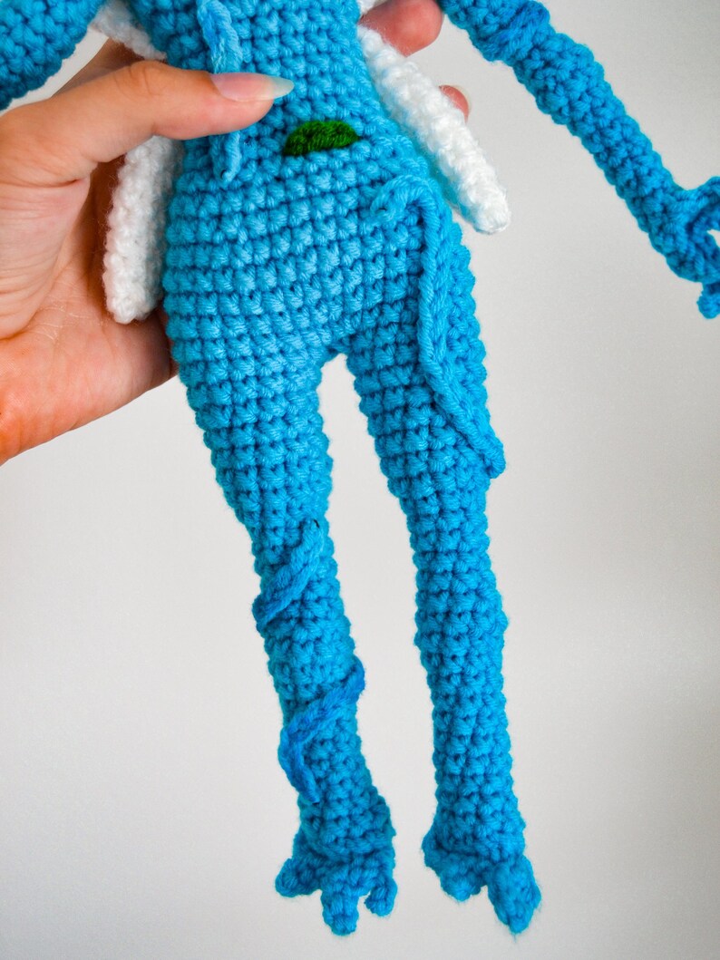 May include: A handmade crochet doll featuring a bright blue body with textured stitching. The doll has white wing-like appendages attached to the upper back and a small green accent detail at the waist. The legs are long and slender with blue yarn detailing.