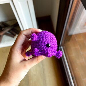 Op de afbeelding: Een kleine, felpaarse octopus gemaakt van gehaakt garen. Het pluche figuurtje heeft een ronde kop, een textuur die is gecre&euml;erd door de steken en verschillende kleine, gekrulde tentakels die vanaf de basis uitsteken. Het heeft &eacute;&eacute;n zichtbaar zwart plastic oog.