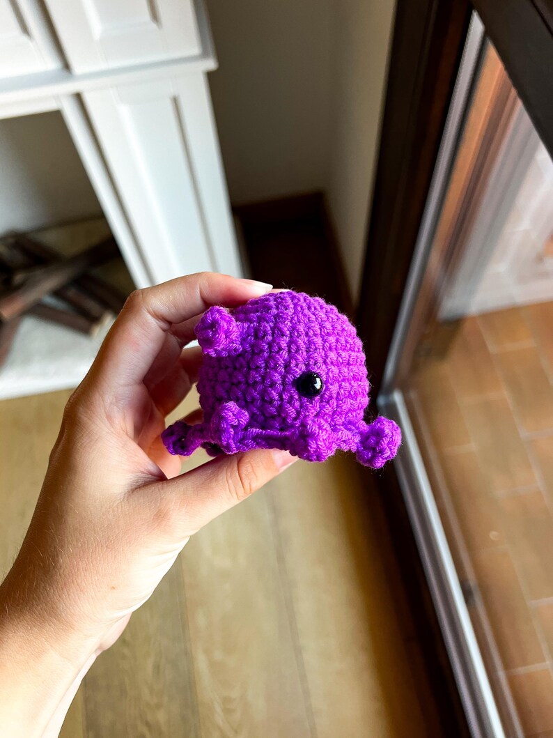 Op de afbeelding: Een kleine, felpaarse octopus gemaakt van gehaakt garen. Het pluche figuurtje heeft een ronde kop, een textuur die is gecre&euml;erd door de steken en verschillende kleine, gekrulde tentakels die vanaf de basis uitsteken. Het heeft &eacute;&eacute;n zichtbaar zwart plastic oog.